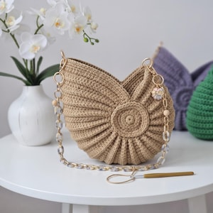 May include: A beige crocheted handbag with a shell-like pleated design, featuring a gold-tone metal chain strap with marbled acrylic links, a small circular charm, and a gold-handled crochet hook resting on the surface in front of the bag.