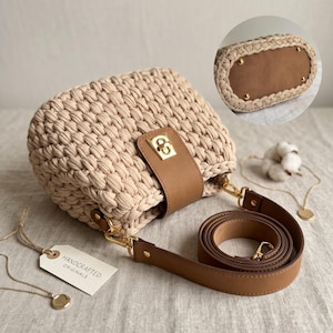 May include: A beige crocheted handbag with a tan faux leather flap featuring a gold-tone twist lock closure. The bag includes a detachable tan faux leather shoulder strap and a tag labelled Handcrafted Originals. An inset shows the brown base.