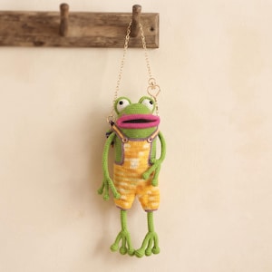 May include: A crochet frog bag featuring a green body with large white and black eyes and a bright pink mouth. The frog wears yellow and white striped overalls with purple trim and is suspended by a thin gold-toned metal chain strap.