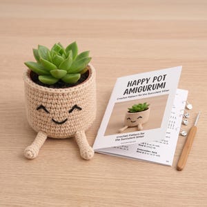 Op de afbeelding: Een beige gehaakte pothoes met een lachend gezicht en twee bungelende benen, met daarin een groene vetplant. Daarnaast ligt een gedrukt instructieboekje genaamd Happy Pot Amigurumi, een bamboe haaknaald en diverse metalen steekmarkeerders.