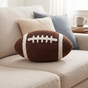 May include: A brown crochet football-shaped cushion featuring white stitched laces and two vertical white stripes on each side. The textured yarn construction mimics the appearance of a classic American football in a soft, plush, decorative format.