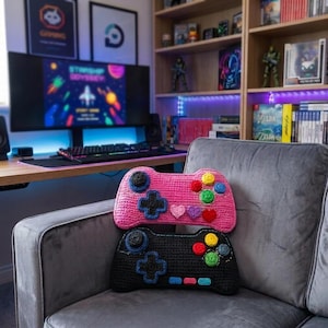 以下が含まれることがあります： ビデオゲームコントローラーの形をした手作りのクロッシェ編みクッション2つ。上のクッションは鮮やかなピンク色で、黒いジョイスティック、黒い十字キー、ハート型を含むカラフルなボタンが付いています。下のクッションは黒色で、青いジョイスティックと十字キー、黄色、緑色、青色の丸いボタンが付いています。