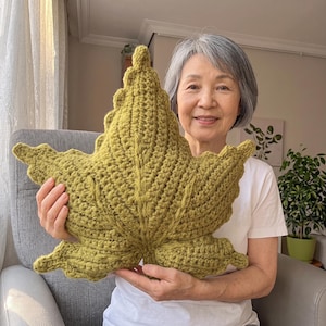 Può includere: Un grande cuscino a forma di foglia color verde oliva, realizzato con un punto all'uncinetto testurizzato. Il cuscino presenta una nervatura centrale distinta e diversi lobi appuntiti, che imitano l'aspetto di una foglia d'acero naturale.