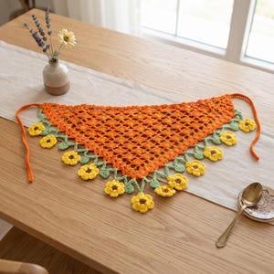 Puede incluir: Un pañuelo triangular de ganchillo con un patrón naranja de tejido abierto. El borde inferior está decorado con un ribete de enredadera verde festoneado con pequeñas flores de color amarillo brillante con centros marrones. Incluye lazos naranjas.
