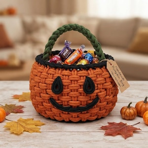 Pode incluir: Uma cesta redonda tecida de cor laranja em forma de abóbora com um rosto preto costurado, uma alça trançada verde e uma etiqueta de papel castanho com a inscrição "Handmade Pumpkin Bag - Happy Halloween". A cesta está cheia de doces embalados.