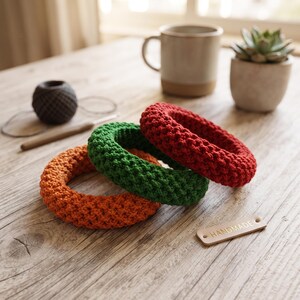 May include: Three circular crochet rings in orange, green, and red, featuring a textured stitch pattern. A small rectangular leather-like tag with the word HANDMADE embossed in gold is placed near the rings, which are arranged on a wooden surface.