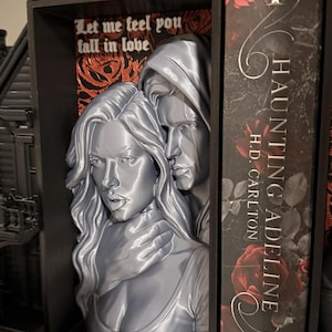 Op de afbeelding: Een zwarte rechthoekige boekensteun met een zilverkleurig 3D-reliëf van twee figuren, de een achter de ander, met de tekst Let me feel you fall in love op een achtergrond van rode rozen. De boekensteun staat naast een boek genaamd Haunting Adeline.