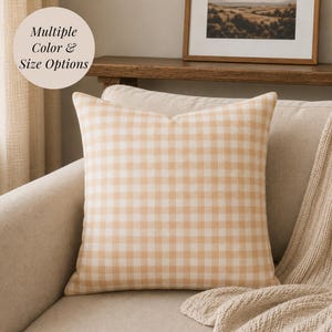 May include: A square throw pillow featuring a light beige and white gingham check pattern. The pillow has a soft, woven texture and is displayed on a neutral-toned sofa. A circular graphic in the upper left corner contains the text Multiple Color & Size Options.