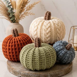 May include: A collection of four handcrafted crochet pumpkins in varying sizes and colors, including a large cream cable-knit pumpkin, a medium orange waffle-stitch pumpkin, a medium sage green ribbed pumpkin, and a small blue textured pumpkin.