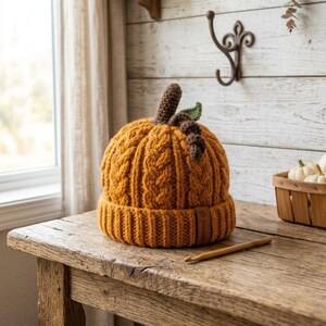 May include: A pumpkin-shaped beanie hat knitted in orange yarn with a cable knit pattern. The hat features a brown crocheted stem and a small green leaf on top. A small rectangular tag labeled "HANDMADE" is attached to the ribbed brim.