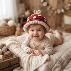 May include: A crochet hat shaped like a mushroom with a deep red cap, white circular spots, and a cream-colored brim. The hat features a small loop at the top and is displayed alongside a wooden tag labeled HANDMADE MUSHROOM CAP.