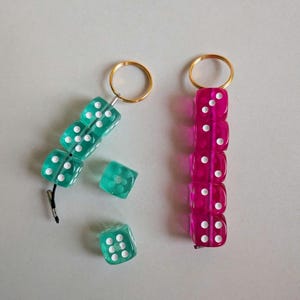 Pode incluir: Dois porta-chaves com dados translúcidos. Um tem três dados azul-petróleo presos a um anel dourado, com dois dados soltos ao lado. O outro consiste em cinco dados cor-de-rosa translúcidos enfiados num anel dourado.