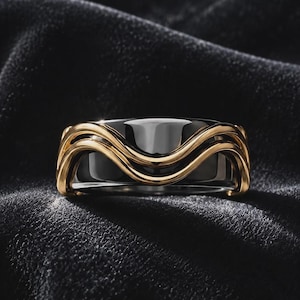 May include: A ring featuring a polished black band with a prominent, raised gold-toned metal overlay that forms a continuous, flowing wave pattern around the circumference, creating a striking contrast between the dark and metallic surfaces.