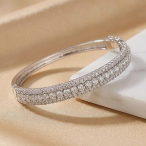 Puede incluir: Una pulsera rígida articulada de tono plateado con tres filas paralelas de pequeñas piedras redondas transparentes facetadas en toda la superficie exterior. La pulsera tiene un mecanismo de cierre seguro visible en la parte superior derecha.