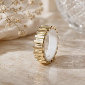 Puede incluir: Un anillo que presenta un exterior de tono dorado con una serie de segmentos rectangulares verticales dispuestos en fila alrededor de la banda. El interior del anillo es liso y blanco, contrastando con la superficie exterior de metal dorado.