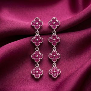 May include: A pair of drop earrings featuring four vertically aligned clover-shaped motifs. Each clover is set with vibrant pink stones and framed by a metallic border. The earrings have a dangling design with a consistent pattern of four segments.