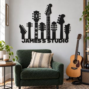 May include: A black metal wall art piece featuring silhouettes of seven different guitar headstocks arranged in a row, with the text "JAMES'S STUDIO" cut out in bold, capital letters directly beneath the guitar necks.