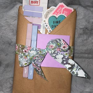 May include: A brown paper envelope containing stationery items including two pastel pens, a sheet of purple sticky notes, and several stickers with text such as Tab It Like It's HOT and FAKE DATE. The package is tied with a floral patterned ribbon.