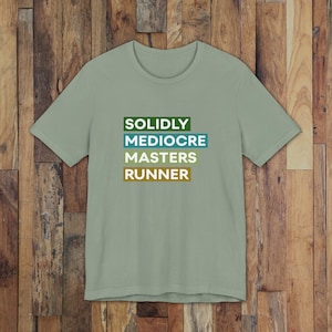 May include: A sage green short-sleeve t-shirt featuring a graphic print with four stacked rectangular blocks in dark green, teal, light green, and brown. The text reads SOLIDLY in the first block, MEDIOCRE in the second, MASTERS in the third, and RUNNER in the fourth.