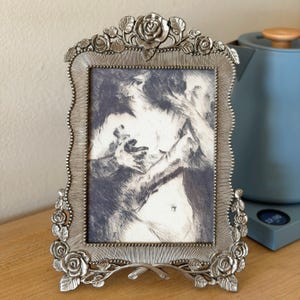Peut inclure: Un cadre photo rectangulaire en métal argenté doté d'une bordure ornée de roses et de feuilles sculptées. Le cadre contient un croquis en noir et blanc représentant une silhouette aux bras levés, sur un fond texturé et rayonnant.