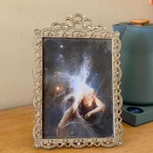 Peut inclure: Un cadre photo rectangulaire avec une bordure métallique argentée ornée de volutes complexes et de petits accents en forme de perles. Le cadre contient une impression représentant une silhouette flottant dans un espace cosmique sombre.