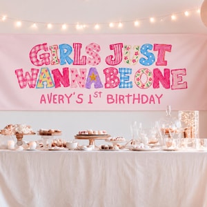 以下が含まれることがあります： GIRLS JUST WANNA BE ONEおよびAVERY'S 1ST BIRTHDAYというカラフルな文字が書かれたライトピンクの長方形のバナー。バナーは白いテーブルクロスがかけられたテーブルの上に飾られており、テーブルの上にはデザートスタンドやスイーツが並んでいます。