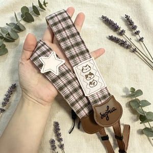 May include: A camera strap featuring a brown, white, and pink plaid pattern. It includes a white star-shaped patch, a rectangular patch with three embroidered cat faces, and a brown leather-like tab with the name Sophia printed in black script.