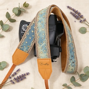 May include: A camera strap featuring a blue fabric pattern with gold stars, tan leather end tabs, and a brown woven backing. One leather tab is personalised with the name Bradley in black script. The strap is attached to a black camera body.