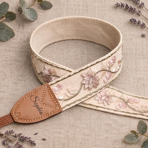 May include: A beige camera strap featuring an embroidered floral pattern in shades of pink and green. The strap includes a tan leather end tab with the name Sophia embossed in black text, finished with visible stitching along the leather edges.