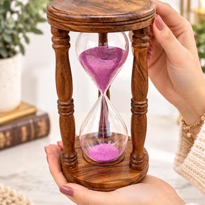 Puede incluir: Un reloj de arena con un marco de madera marrón oscuro y cuatro pilares torneados que conectan una base y una parte superior circulares. El bulbo de vidrio transparente contiene arena púrpura vibrante que fluye de la cámara superior a la inferior.