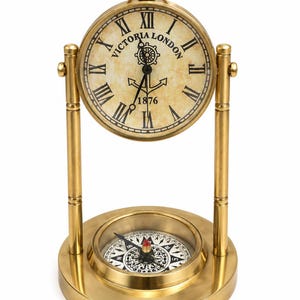 May include: A brass-colored table clock with a round face featuring Roman numerals, the text "VICTORIA LONDON", a ship's wheel, and an anchor graphic. The clock is suspended between two vertical posts above a circular base containing a magnetic compass.