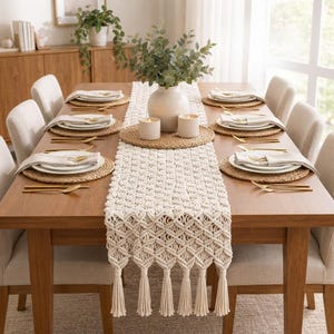 May include: A cream-colored macrame table runner with a geometric knot pattern and long fringe tassels, draped down the center of a wooden dining table set with white plates, gold cutlery, woven placemats, and a central vase with greenery.