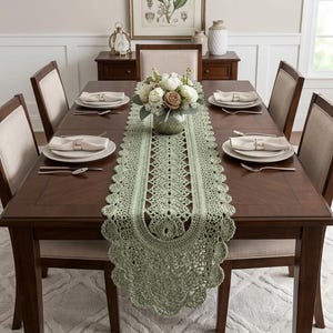 May include: A sage green crocheted table runner with an intricate openwork pattern and scalloped edges, centered on a dark wood dining table. The runner features a central floral arrangement in a glass vase, surrounded by four place settings.