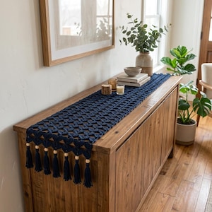 May include: A navy blue crochet table runner featuring a repeating star-like pattern and a decorative edge finished with hanging dark blue tassels and light-colored wooden beads, draped across the top of a wooden cabinet.