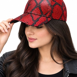 May include: A baseball cap featuring a vibrant red and black snakeskin-like textured pattern. The cap has a structured crown, a curved brim, and a small button at the top center. The material displays a repeating scale-like design throughout.