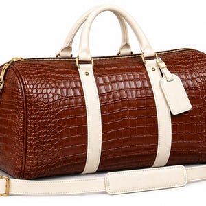 Peut inclure: Un sac de voyage marron avec un motif texturé en peau de crocodile, doté de poignées couleur crème, d'une bandoulière amovible assortie, de ferrures en métal doré et d'une petite étiquette à bagage couleur crème sur la poignée.