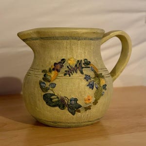 Puede incluir: Una jarra de cerámica de color amarillo claro con una superficie texturizada y un asa. La parte frontal presenta un diseño de corona en relieve compuesto por flores, hojas y una mariposa pintadas en tonos azul, morado, naranja y verde.