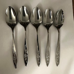 Könnte beinhalten: Ein Set aus fünf silberfarbenen Metalllöffeln, die in einer Reihe angeordnet sind. Jeder Löffel hat eine polierte, reflektierende Laffe und einen schlanken Stiel, der nahe der Basis mit einem Blumenmuster verziert ist.