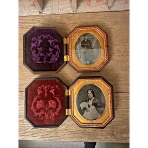 May include: Two open octagonal cases containing vintage daguerreotype portraits. The top case features a purple velvet interior with a gold frame holding a portrait of a person. The bottom case has a red velvet interior and a gold frame with a portrait.