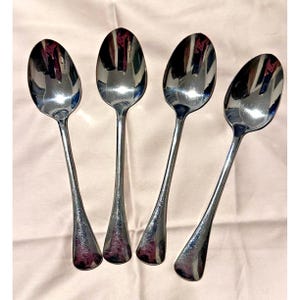 May include: A set of four polished silver-tone metal spoons arranged in a row. Each spoon features a smooth, reflective oval bowl and a slender, slightly flared handle. The metallic surface shows clear reflections of the surrounding environment.