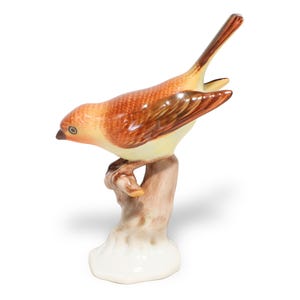 Könnte beinhalten: Eine Keramik-Vogel-Figur mit glänzender Oberfläche, rotbraunem Kopf und Flügeln, hellgelber Brust und dunkelbraunem Schwanz. Der Vogel sitzt auf einem strukturierten, hellbraunen Baumstumpf, der in einen glatten, weißen Sockel übergeht.