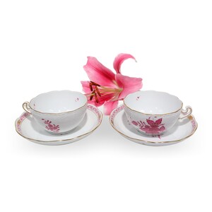 May include: Two white porcelain teacups and matching saucers featuring scalloped edges, gold trim, and a delicate pink floral pattern. A single vibrant pink lily flower is positioned between the two sets, adding a natural accent to the display.
