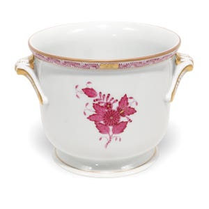 Pode incluir: Um cachepot de porcelana branca com um motivo floral magenta central e duas alças laterais. A borda é decorada com uma faixa magenta e dourada. O recipiente possui uma base circular e um acabamento liso e brilhante.