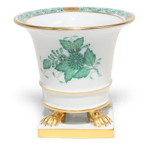 Pode incluir: Um vaso de porcelana branca com um padrão floral verde na frente e uma borda de folhas verdes ao longo da abertura. O vaso tem acabamento dourado, uma abertura alargada e repousa sobre uma base quadrada com quatro pés de garra dourados.