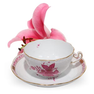 Puede incluir: Un juego de taza de té y platillo de porcelana blanca con borde festoneado, ribete dorado y un patrón floral rosa. Una flor de lirio rosa vibrante se encuentra detrás de la taza, oscureciendo parcialmente la parte posterior del platillo.