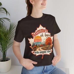 May include: A dark brown short-sleeve t-shirt featuring a large maple leaf graphic. Inside the leaf is a landscape with a sunset, mountains, trees, and a dirt path. The text reads Summer... ok, byeee and hello autumn vibes in a retro font.