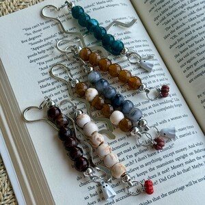 May include: Five metal bookmark clips, each featuring a vertical row of round beads in varying colors including dark green, amber, grey, white, and dark brown. Each bookmark is accented with a small charm shaped like a ghost or a pumpkin.