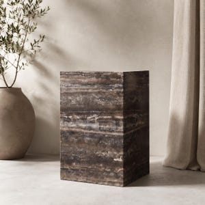 May include: A rectangular pedestal made of dark brown and grey natural stone with visible horizontal veining and a porous, textured surface. The block has sharp, clean edges and a solid, heavy appearance, showcasing the organic patterns of the stone.