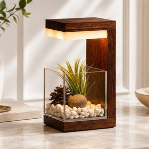 Puede incluir: Una lámpara de madera oscura con un marco en forma de C y una fuente de luz integrada. Bajo la luz, un recipiente cuadrado de acrílico transparente contiene guijarros blancos, una pequeña piña marrón, una piedra beige y una planta de aire.