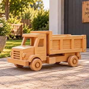 May include: A light-coloured wooden toy dump truck featuring a cab with open windows, a front grille, and a large cargo bed. The vehicle is constructed from smooth, natural-toned wood with visible grain and includes four circular wheels on each side.
